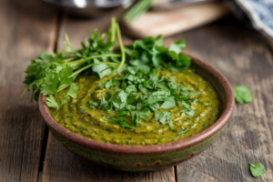 Bowl of vibrant chimichurri sauce with visible herb stems and fresh chopped herbs on a rustic wooden table, with a small cutting board and knife