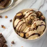 A bowl of peanut butter brownie double cheesecake ice cream with swirls of peanut butter, brownie chunks, and a drizzle of chocolate sauce on a marble background.