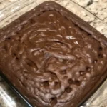 A stack of fudgy cocoa brownies with a gooey center, drizzled with melted chocolate, on a rustic wooden cutting board.