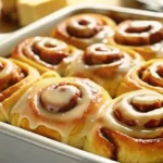 A baking dish filled with fluffy, golden-brown cinnamon rolls drizzled with heavy cream glaze, with a fork lifting one roll to reveal its gooey layers.