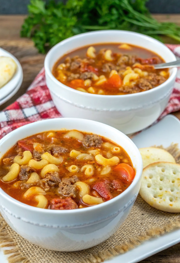 A steaming bowl of goulash soup with beef and tomato macaroni soup garnished with fresh parsley and parmesan cheese, served with crusty bread