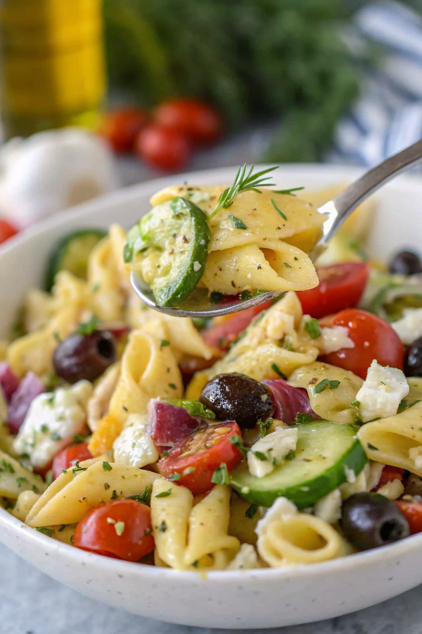 Finished Greek pasta salad in a large white serving bowl, showing colorful rotini pasta mixed with cherry tomatoes, feta cheese, Kalamata olives, cucumber, and fresh herbs, garnished with extra feta and oregano