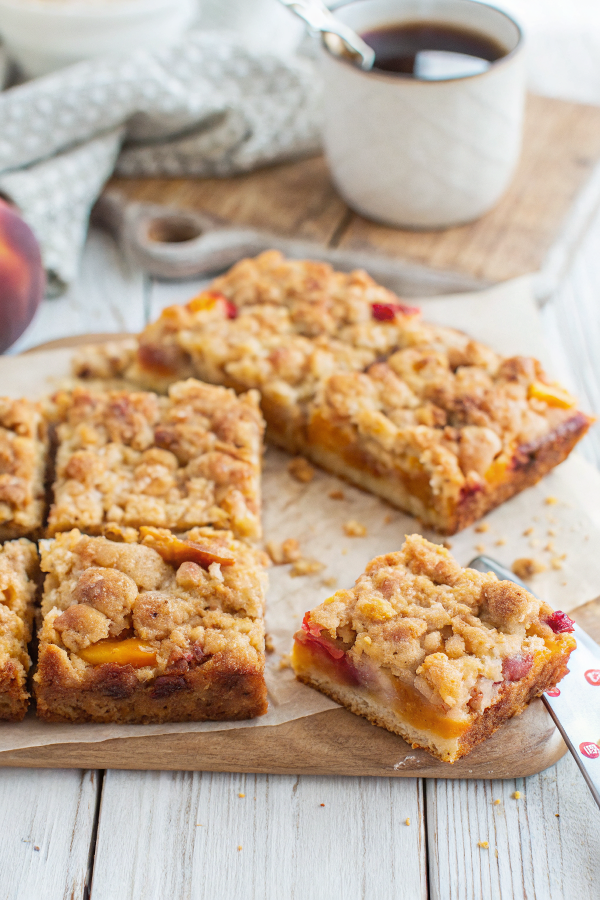 Perfectly sliced peaches and cream crumble bars arranged on rustic wooden board with fresh peach slices and golden lighting